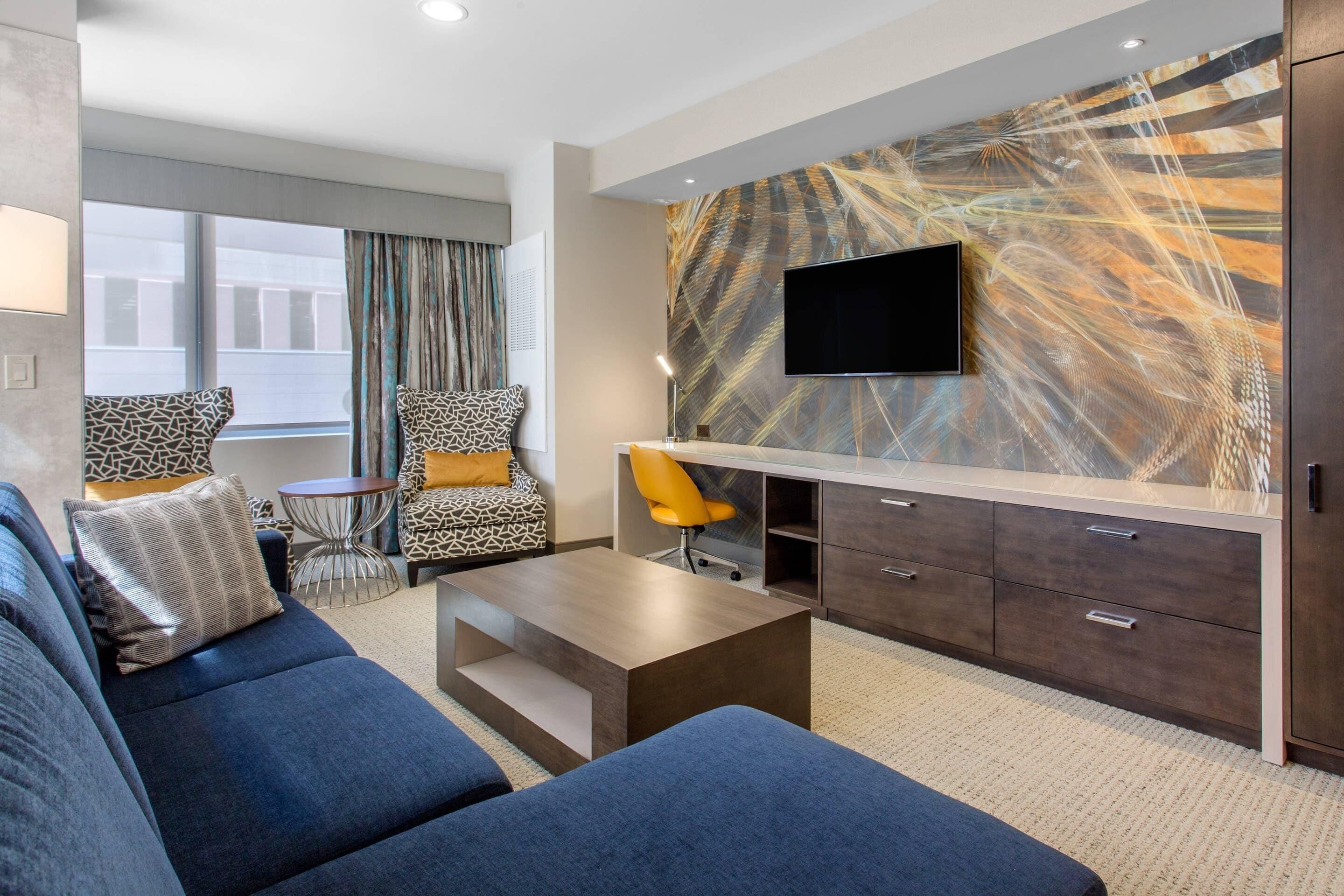 A modern hotel room with a large sofa, two patterned armchairs, a small round table, a TV mounted on a marble wall, and a wooden desk with drawers.