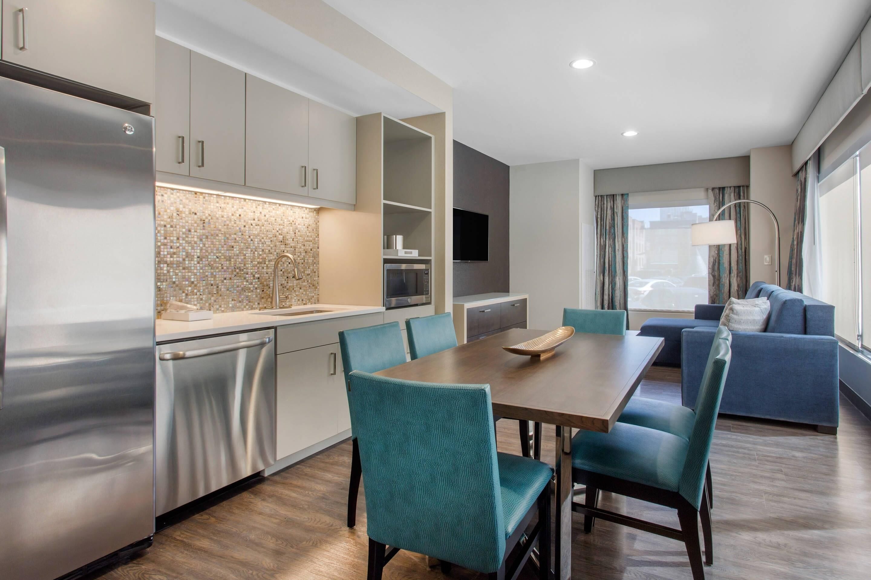 A modern kitchen with a built-in oven, a dining area with teal chairs, and a sofa in the living area.