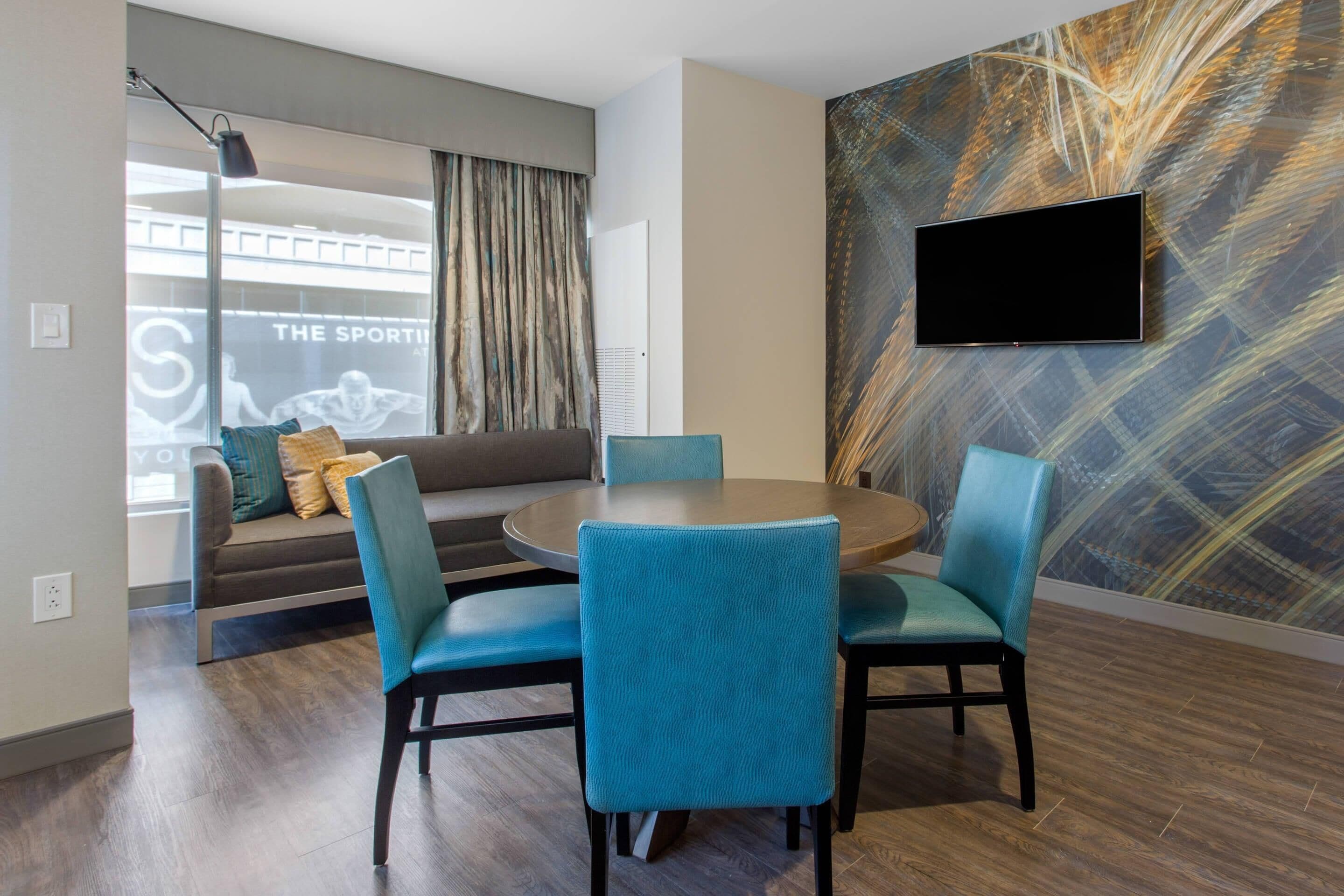 A modern hotel room with a round dining table, two blue chairs, a sofa with cushions, a large abstract wall mural, and a flat-screen TV mounted on the wall.