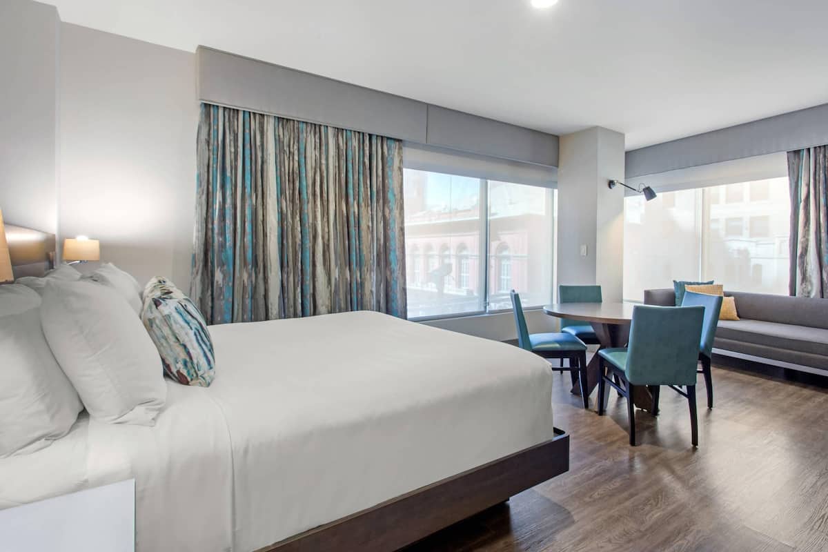 A hotel room with a large bed, a desk, a chair, a sofa, and a view of buildings through the windows.