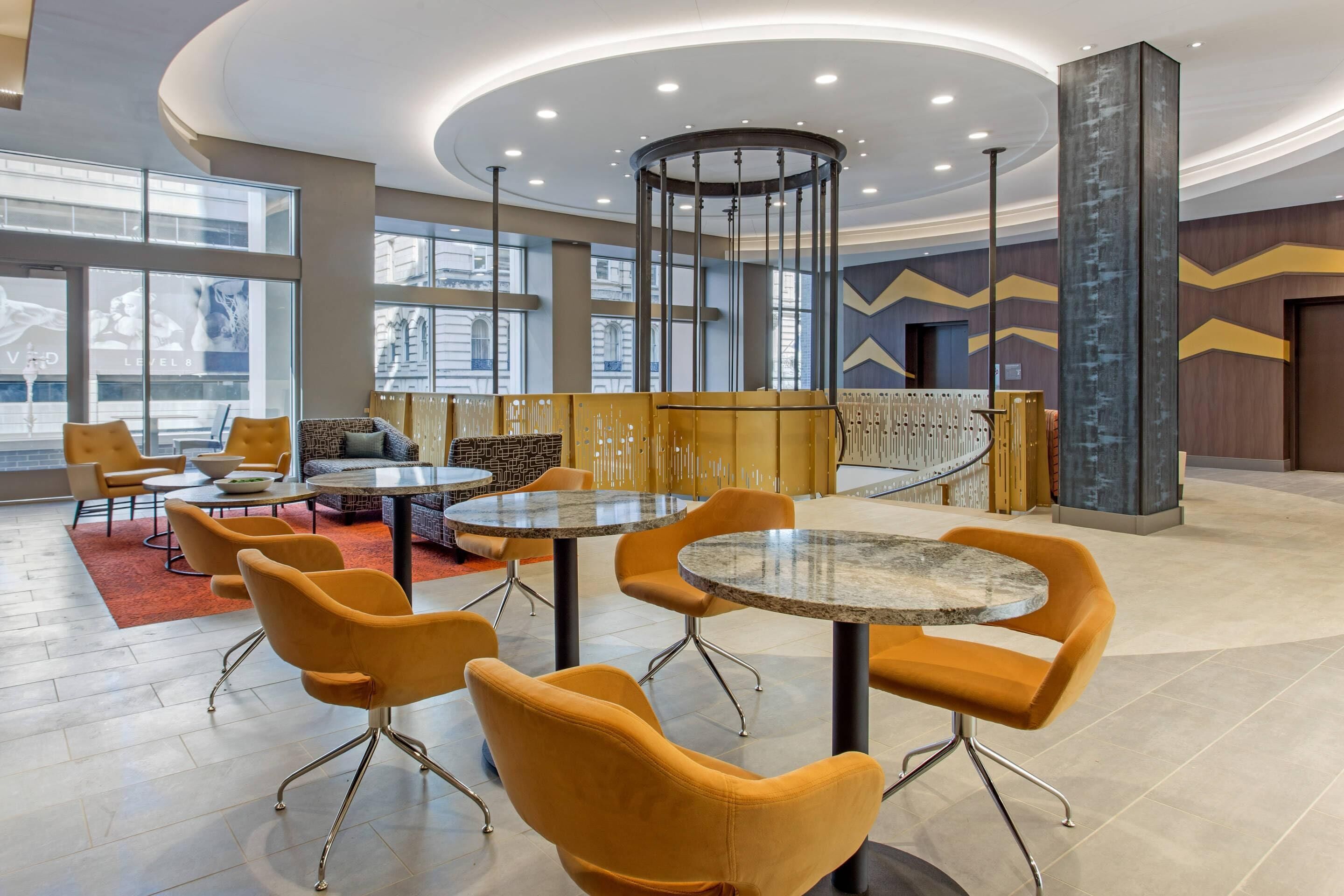 A modern lobby with round tables and orange chairs, large windows, and a central decorative column.