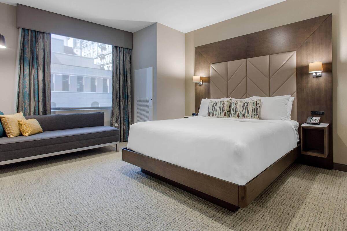 A modern hotel room with a large bed, a sofa, a bedside table with a lamp, and a view of the city through the window.