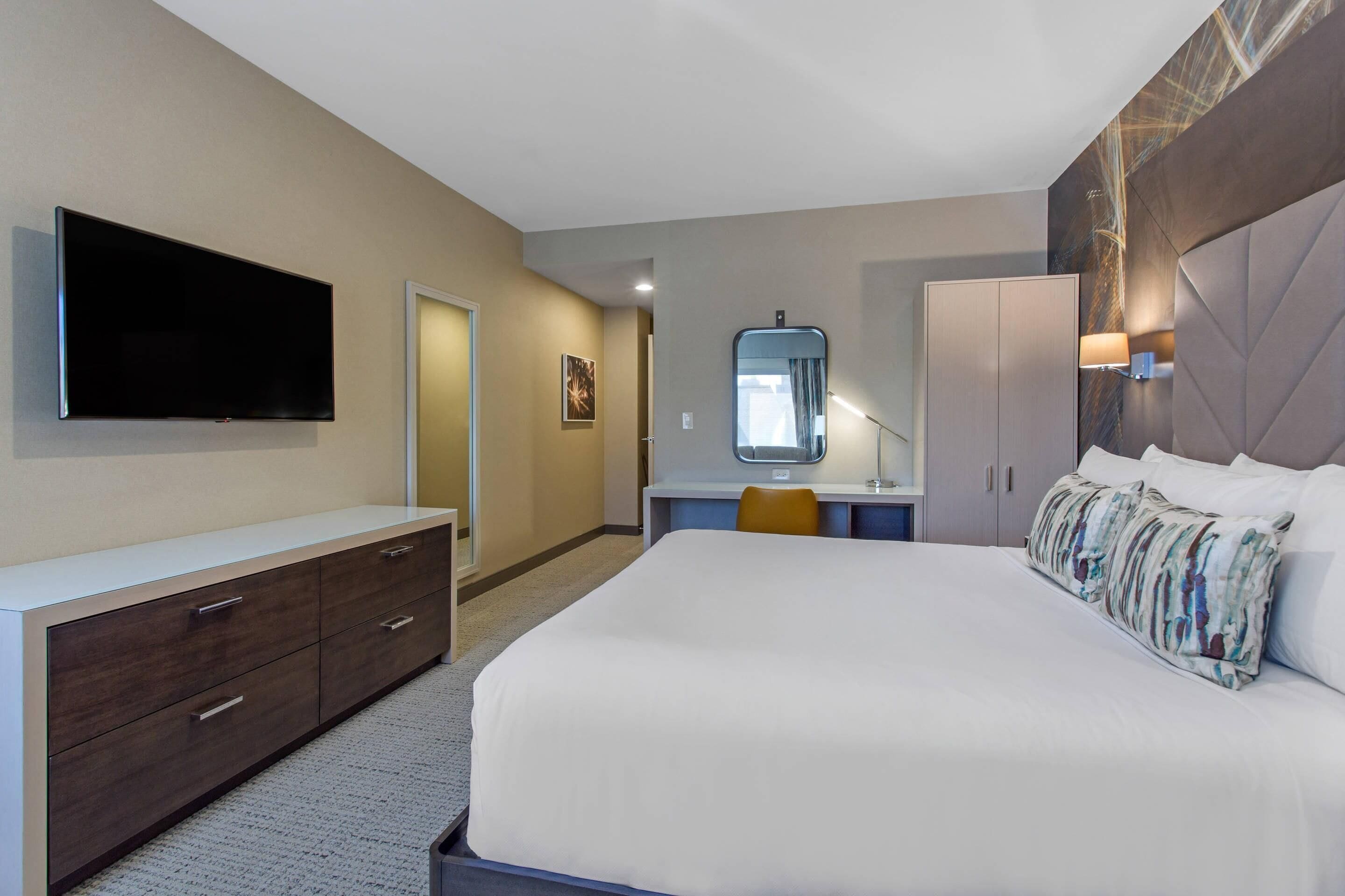 A hotel room with a large bed, a flat-screen TV mounted on the wall, a desk with a chair, and a wardrobe.