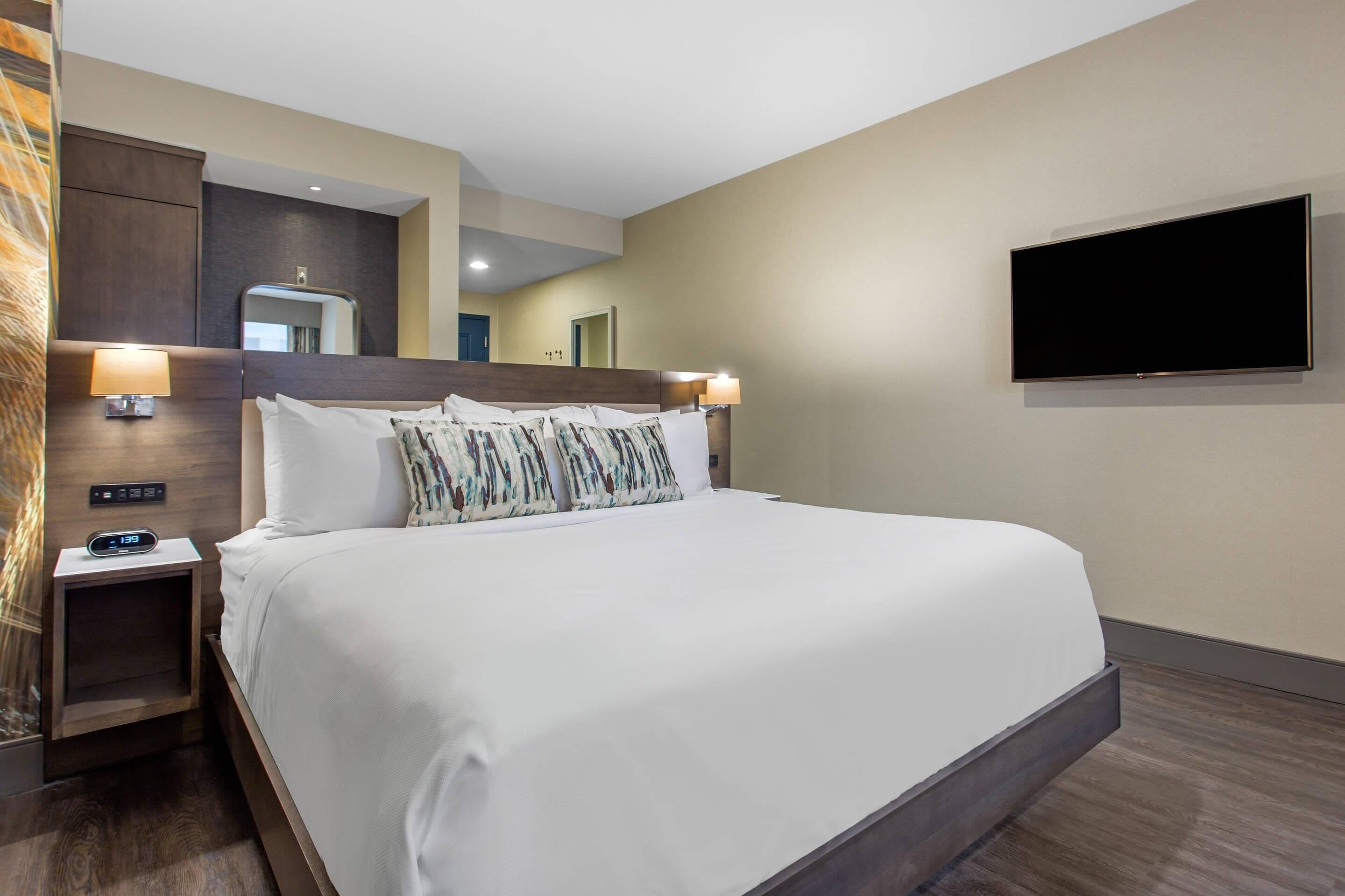Ein Hotelzimmer mit einem großen Bett, zwei Nachttischlampen, einem flachbildfernseher an der Wand und einem Holzkopfstück.