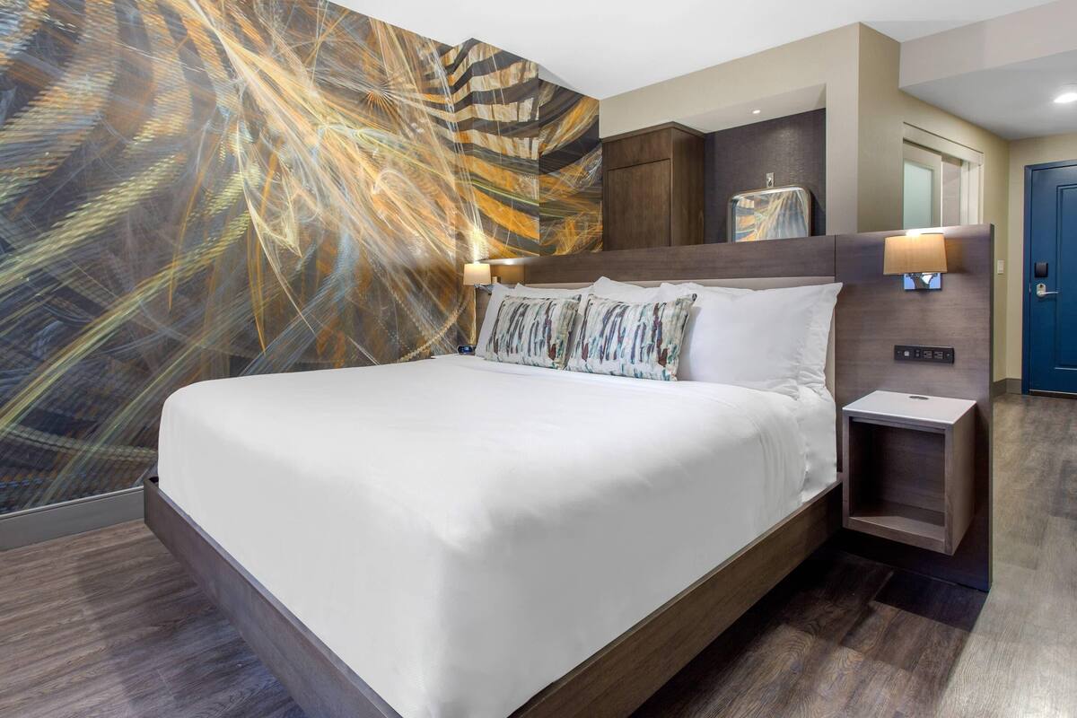 A modern hotel room with a large bed, a nightstand, a wall-mounted lamp, and a decorative abstract wall mural.