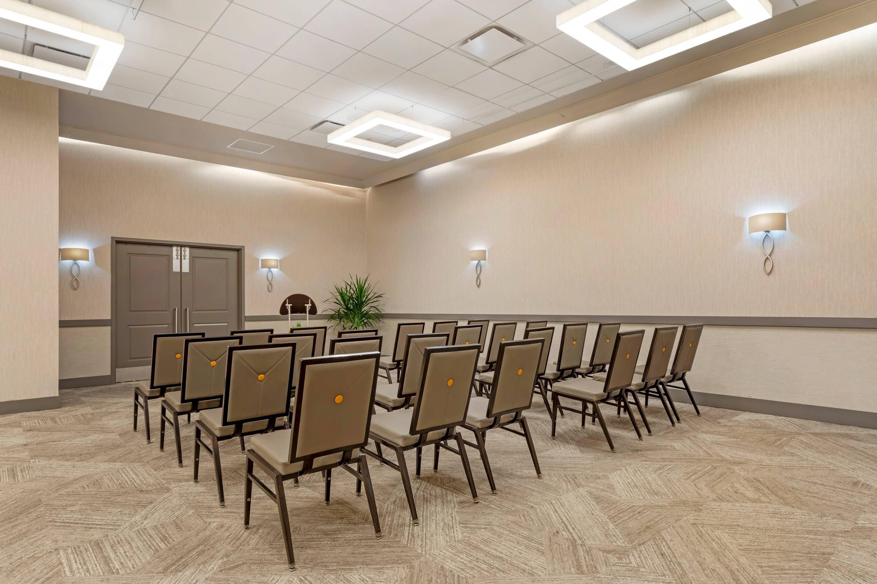 Uma sala de conferências com fileiras de cadeiras e mesas, equipada com luminárias no teto e luminárias fixadas na parede.