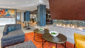 A modern lobby with a sofa, two round tables with bowls, a fire feature, and a dining area in the background.