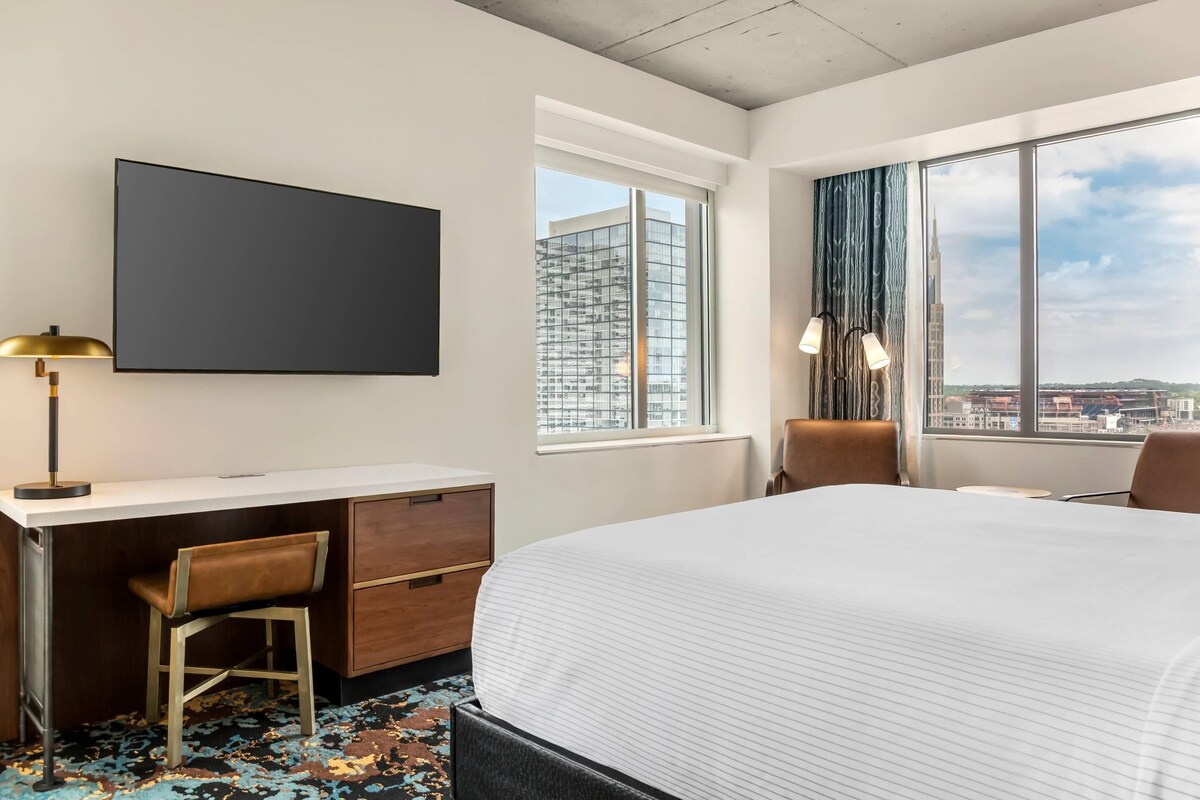 A hotel room with a bed, desk, chair, TV, and large windows offering a city view.