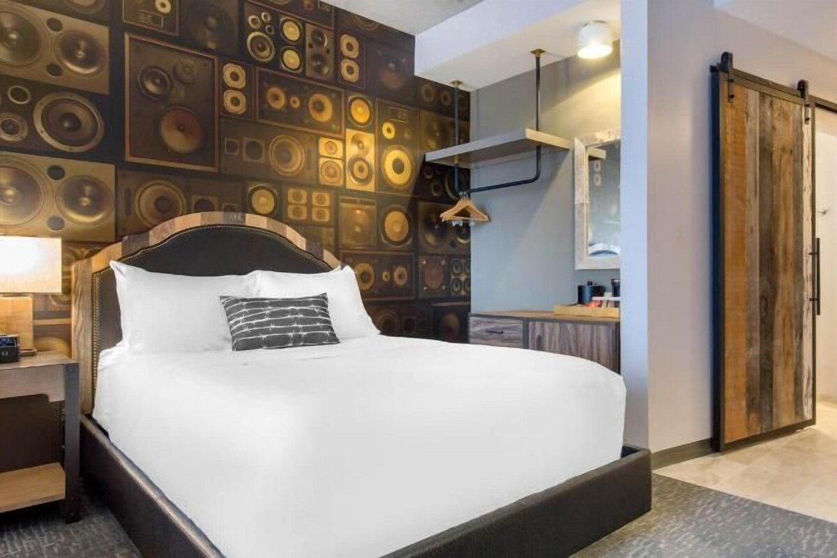 A hotel room with a large bed, a wall of circular patterns, a wooden wardrobe, and a desk with a lamp.