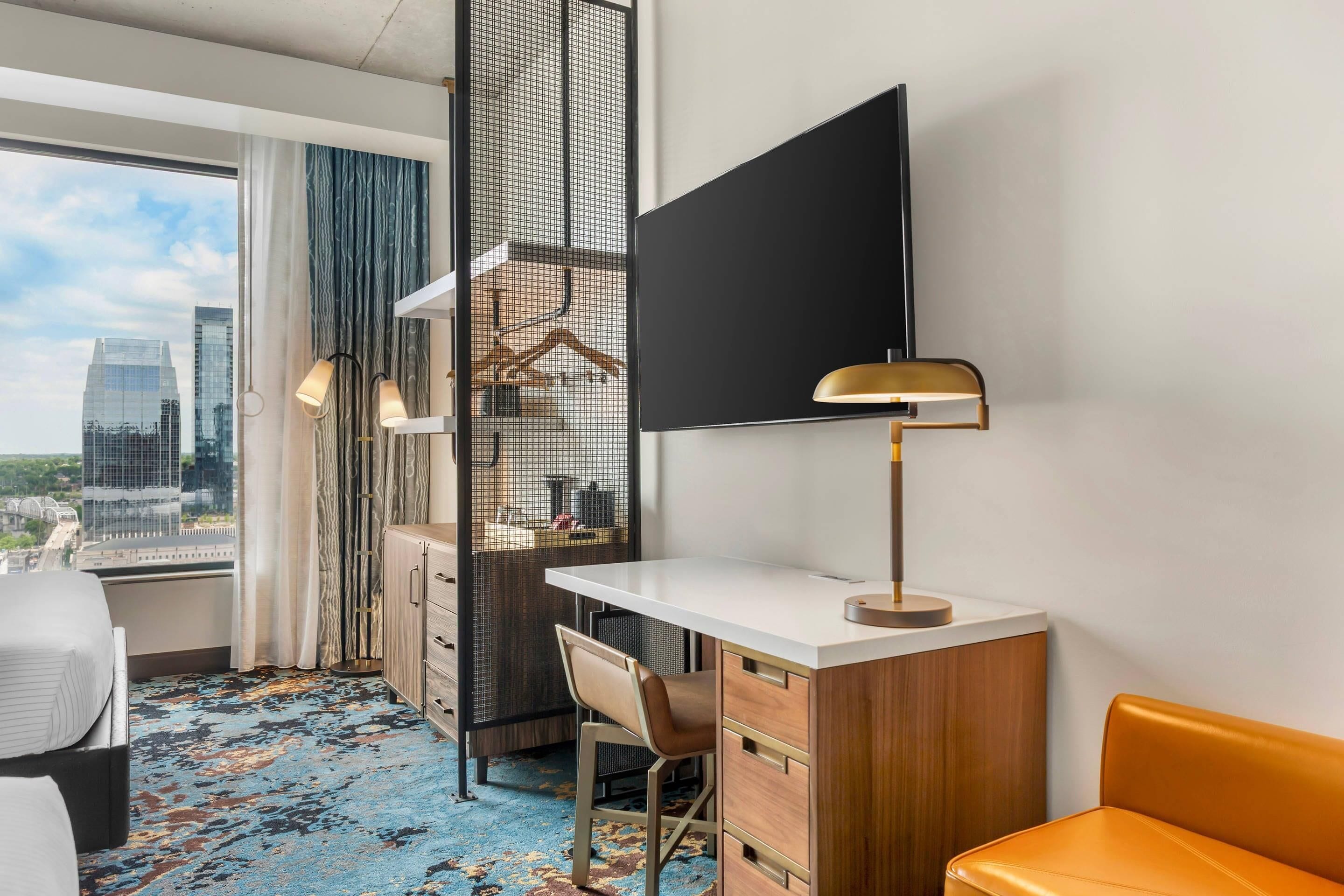 Habitación de hotel con escritorio, silla, cama y un amplio ventanal con vistas a la ciudad.