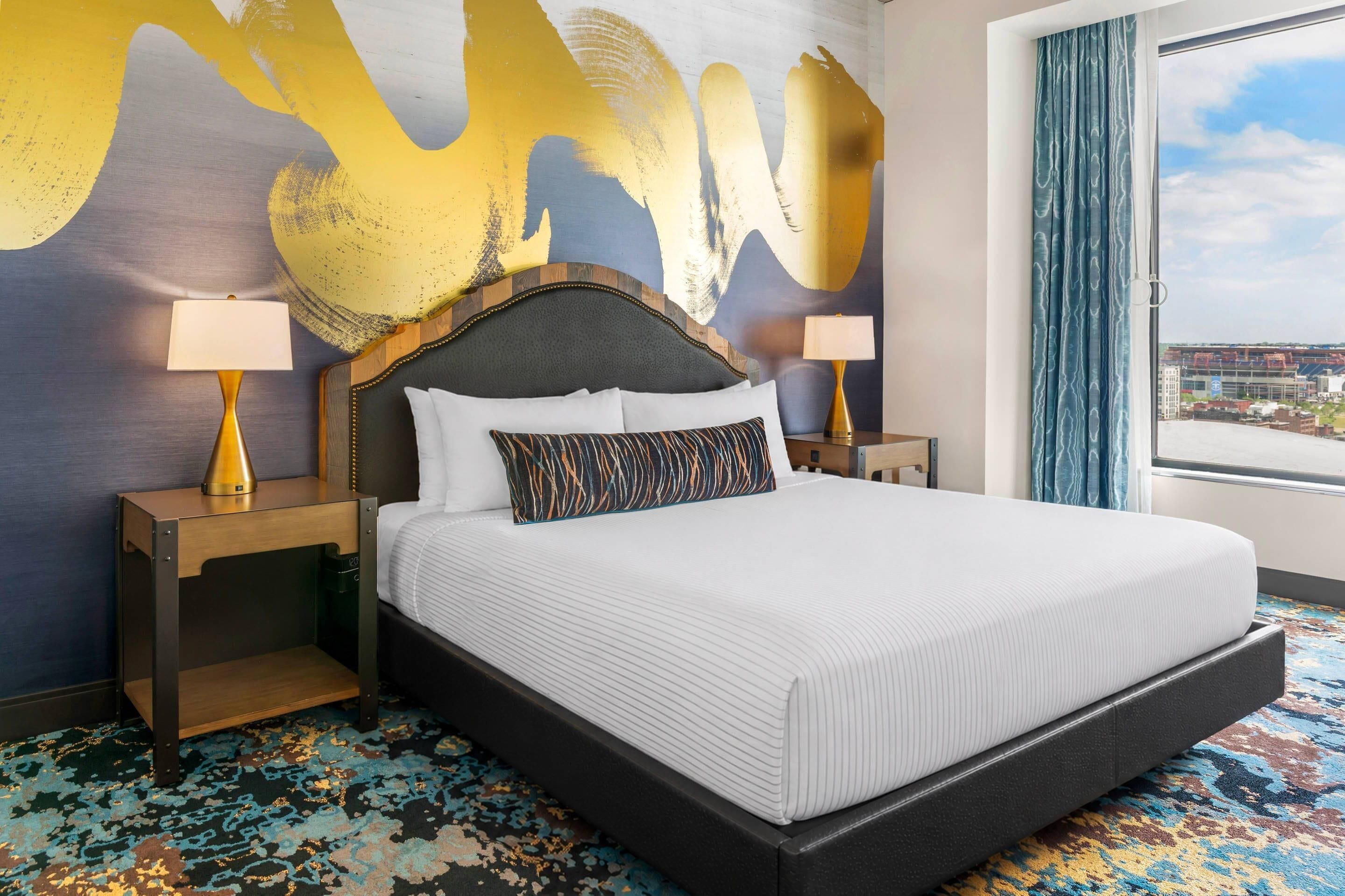 Habitación de hotel con una cama grande, dos mesitas de noche con lámparas, un cabecero moderno y un cuadro abstracto de grandes dimensiones en la pared.