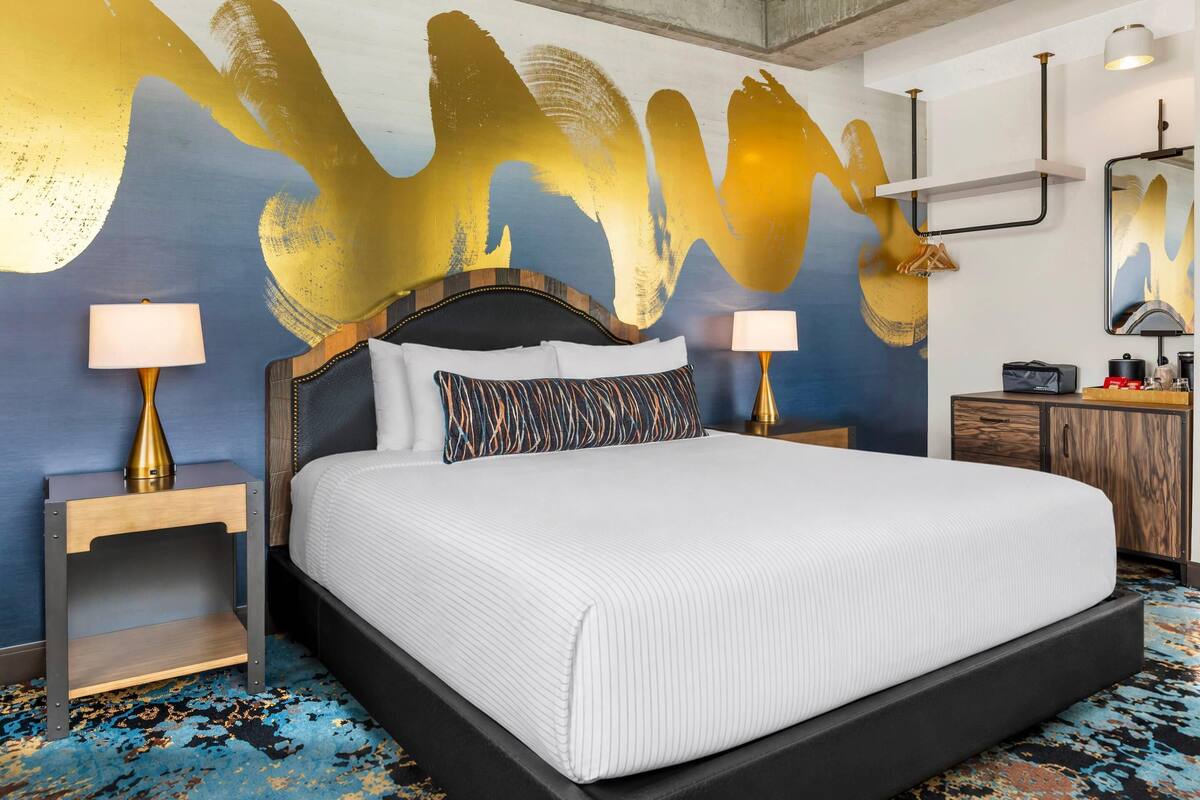 A hotel room with a large bed, two bedside tables with lamps, a wooden headboard, a wall with abstract art, a small cabinet, and a mirror.
