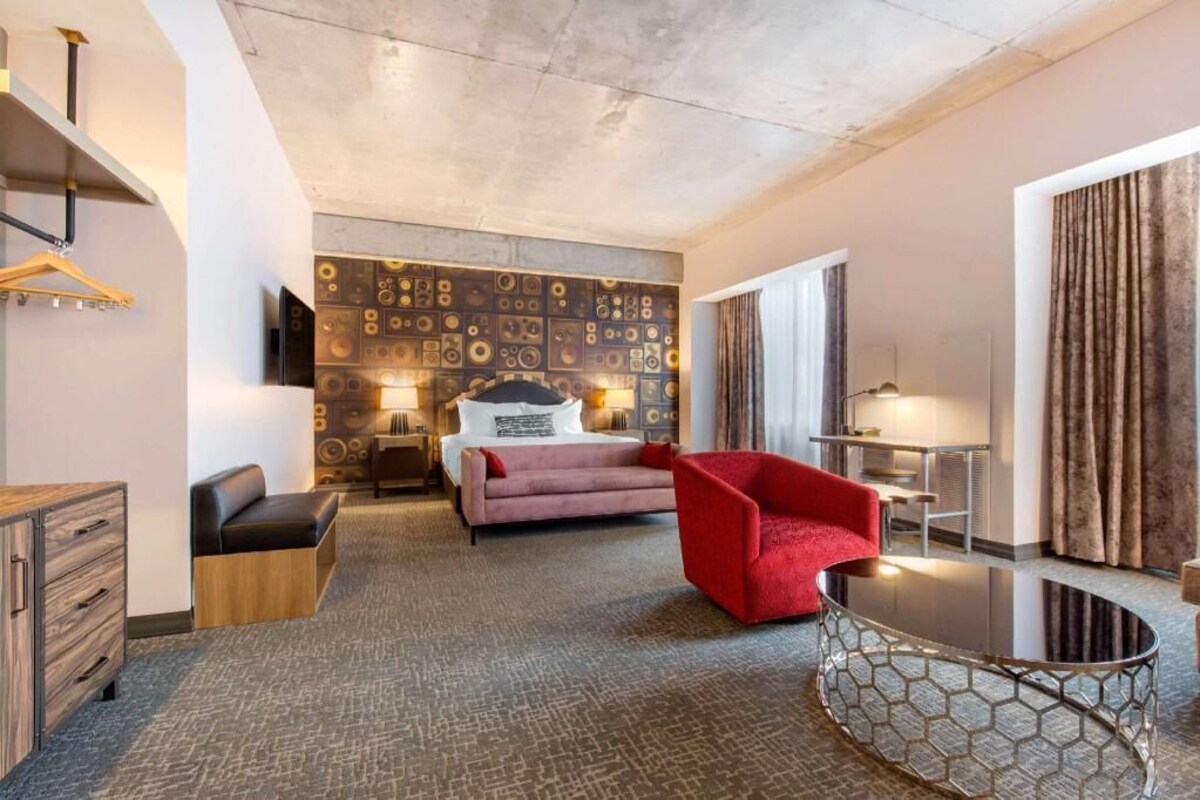 A hotel room with a bed, a red chair, a small table, a dresser, and a wall with a geometric pattern.