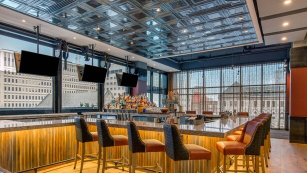 Zona bar moderna con soffitto alto, ampie finestre e bancone con sgabelli.