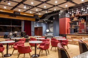 A restaurant with red upholstered chairs, white round tables, a stage with musical equipment, and a bar area with shelves of bottles. - Cambria Hotel Nashville Downtown (Nashville)