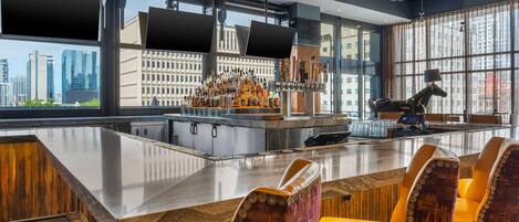 A bar area with a long counter, multiple stools, a bar stocked with bottles, and large windows offering a city view.