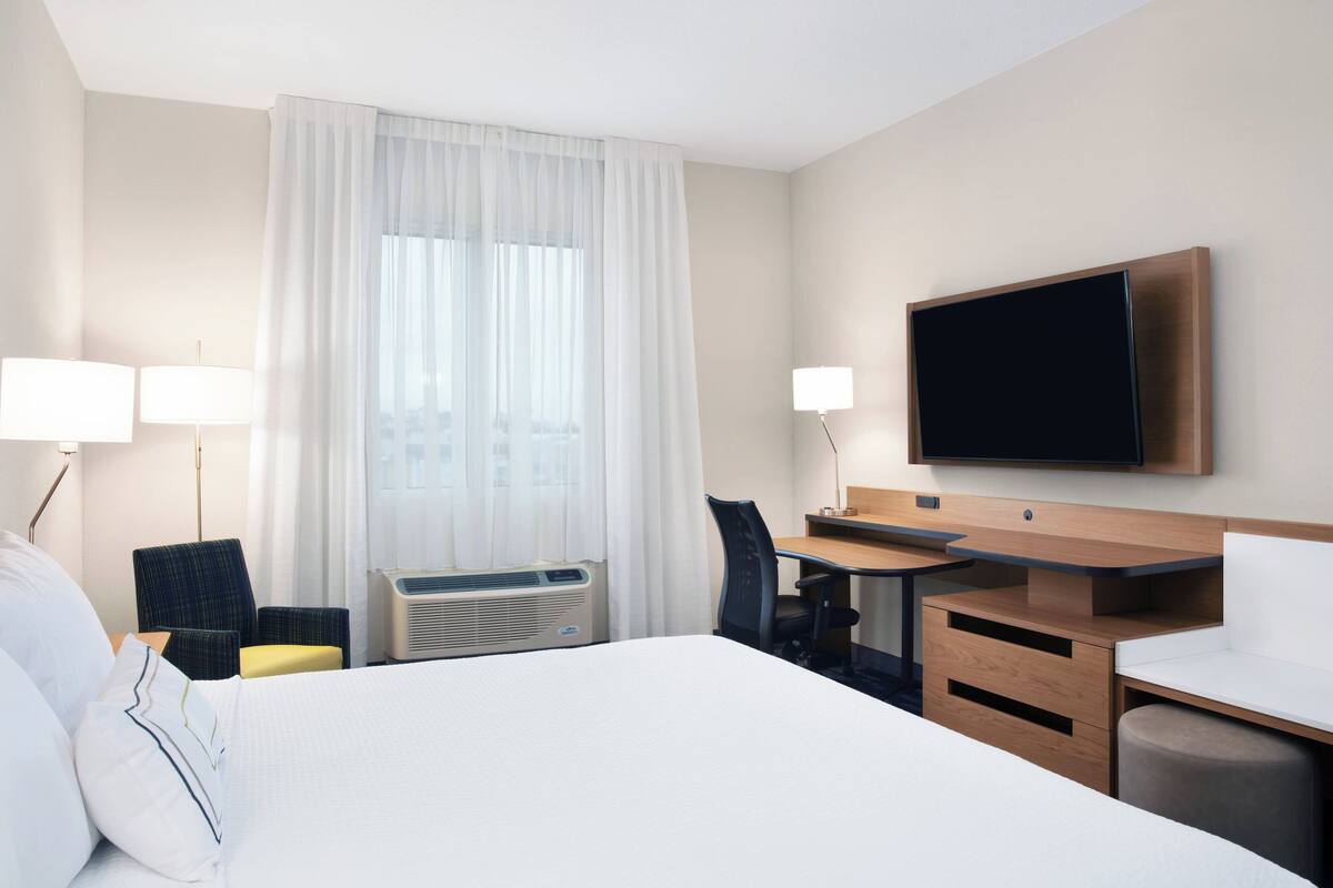A hotel room with a bed, two chairs, a desk, a television, and a window with curtains.