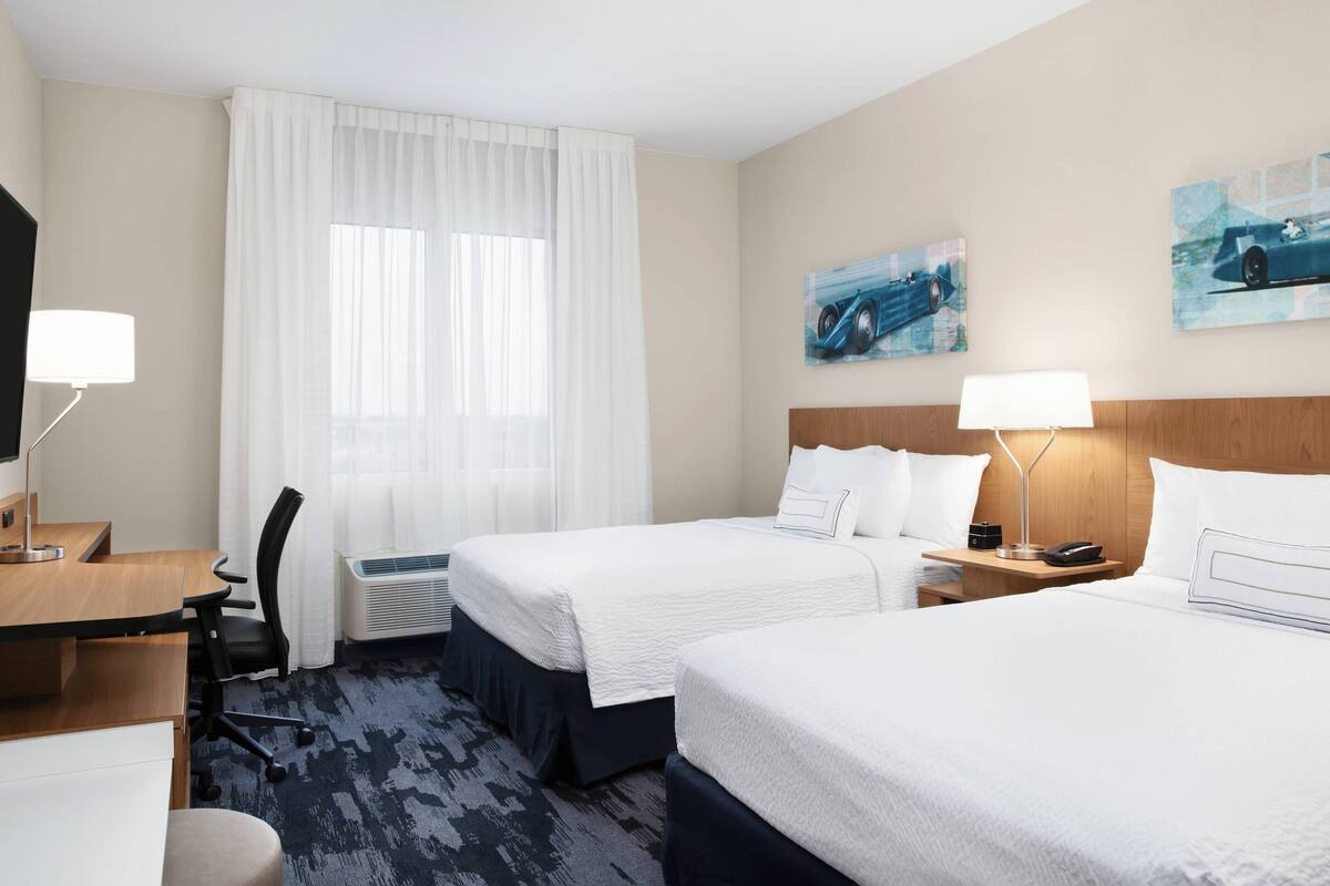 A hotel room with two beds, a desk, a chair, a lamp, and artwork on the walls.