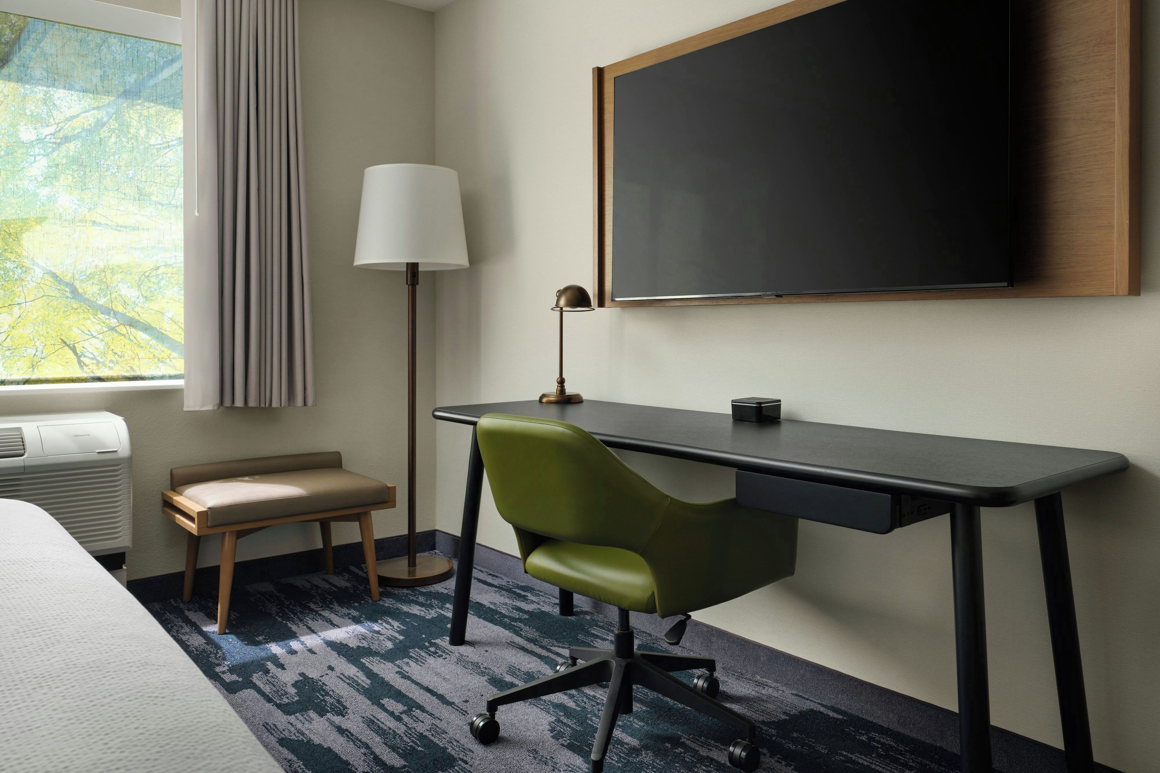 Une chambre d’hôtel avec un lit, un bureau, une chaise, une lampe et une grande fenêtre avec des rideaux.