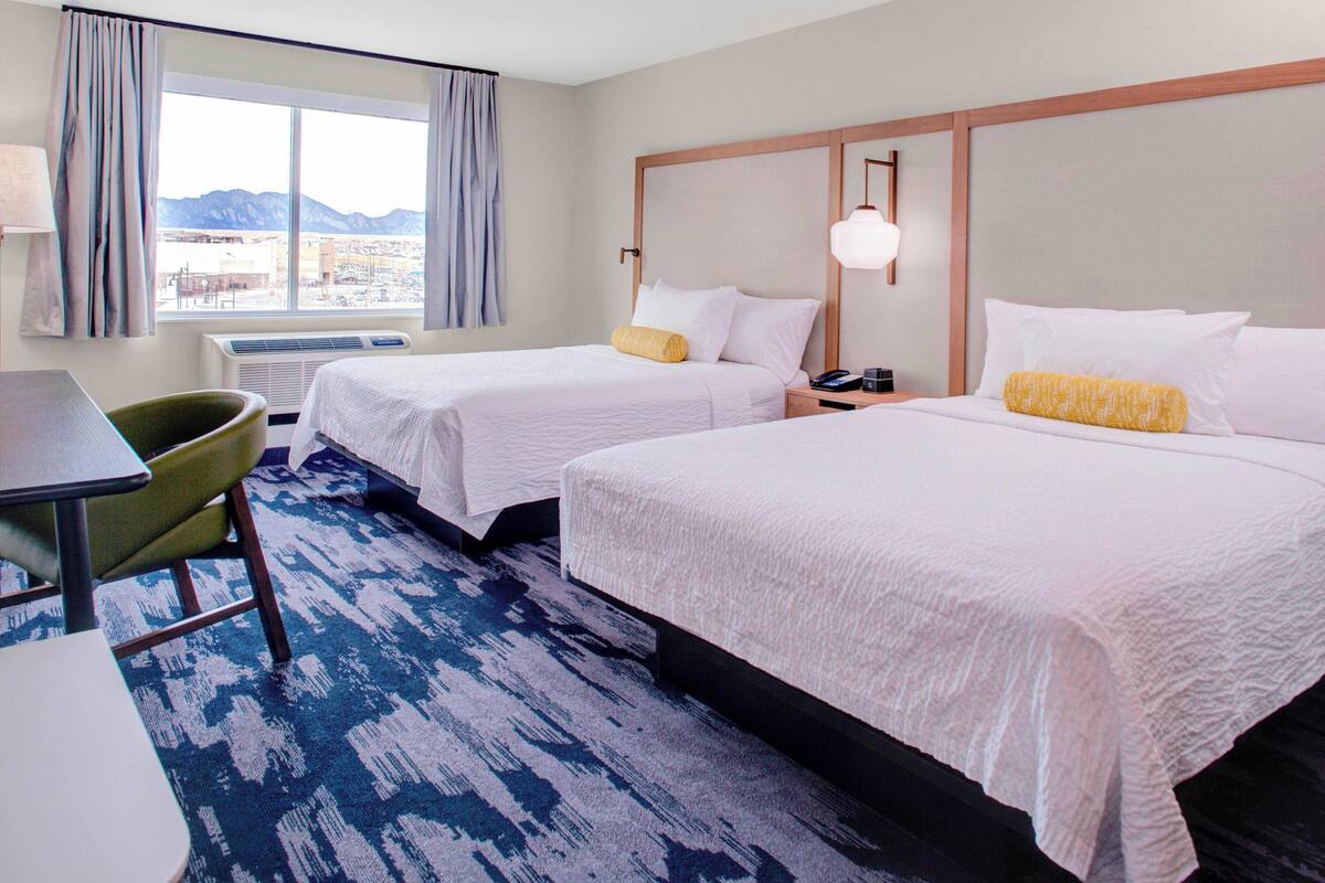 A hotel room with two beds, a desk, a chair, and a view of mountains through the window.