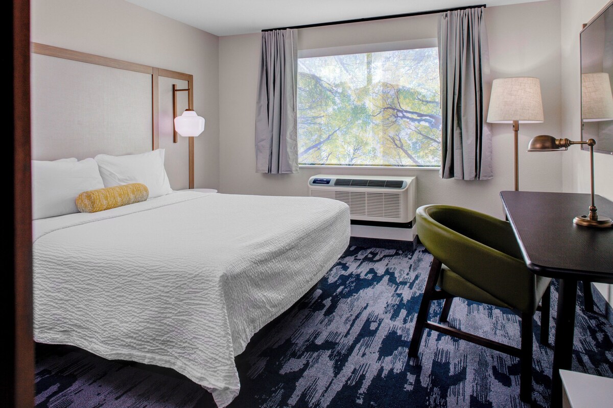 Een hotelkamer met een bed, een bureau, een stoel, een raam met gordijnen en een airconditioner.