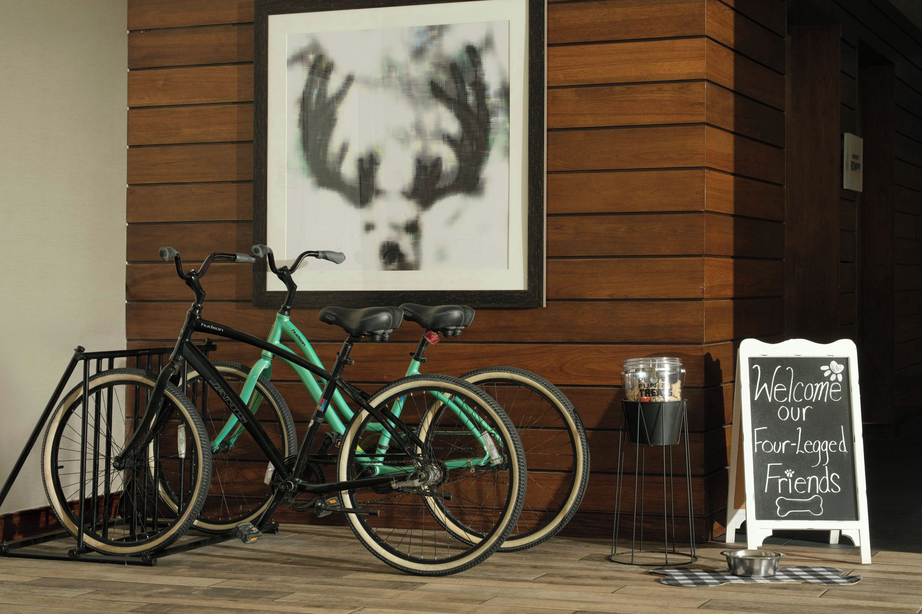 Ein Fahrrad steht in einem Innenraum, mit einer Holzwand und einem gerahmten Bild darüber. Ein Kreidetafel zeigt die Aufschrift 'Willkommen, unsere Vierbeiner'.