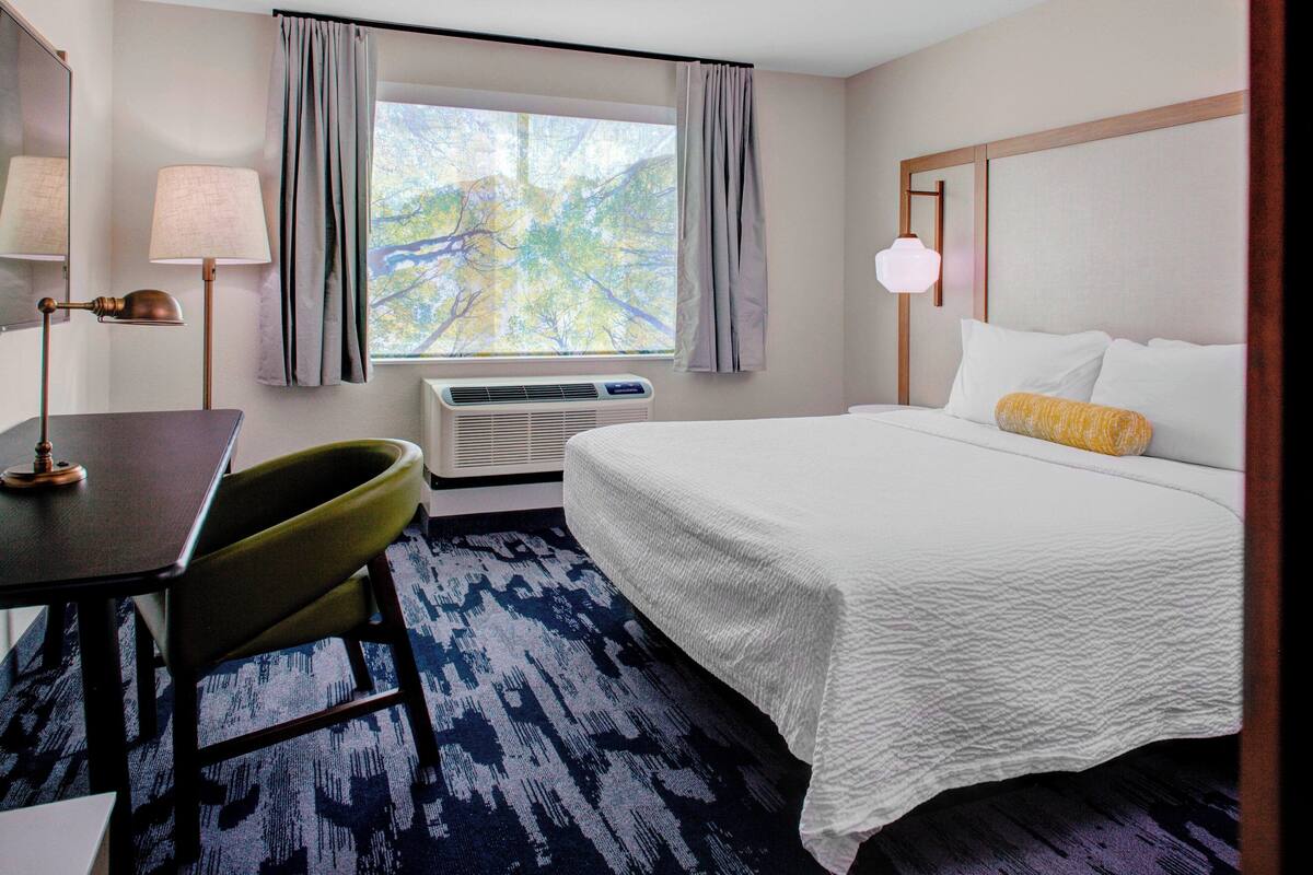 A hotel room with a bed, a desk, a chair, a lamp, a window with curtains, and an air conditioning unit.