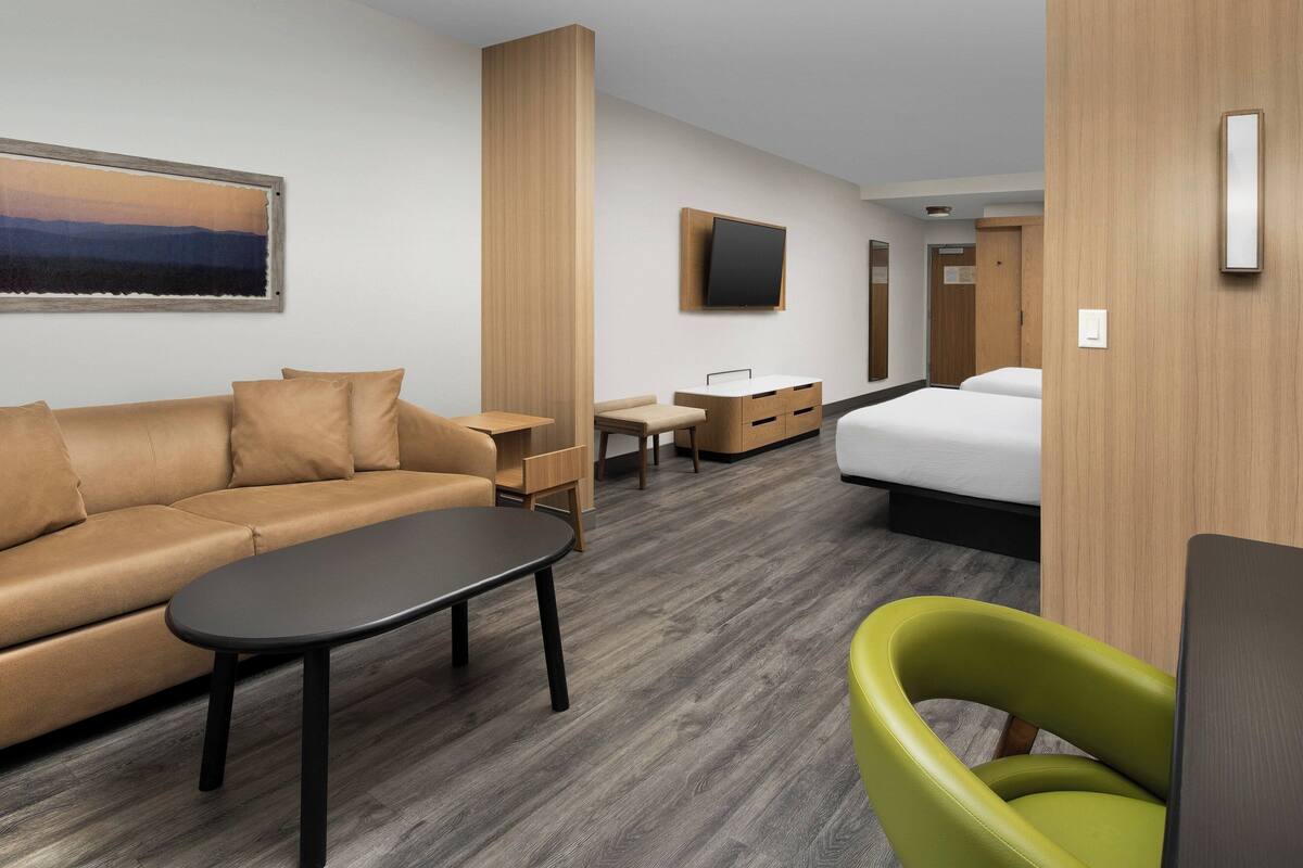 A modern hotel room with a sofa, a small table, a green chair, a bed, and a TV mounted on the wall.