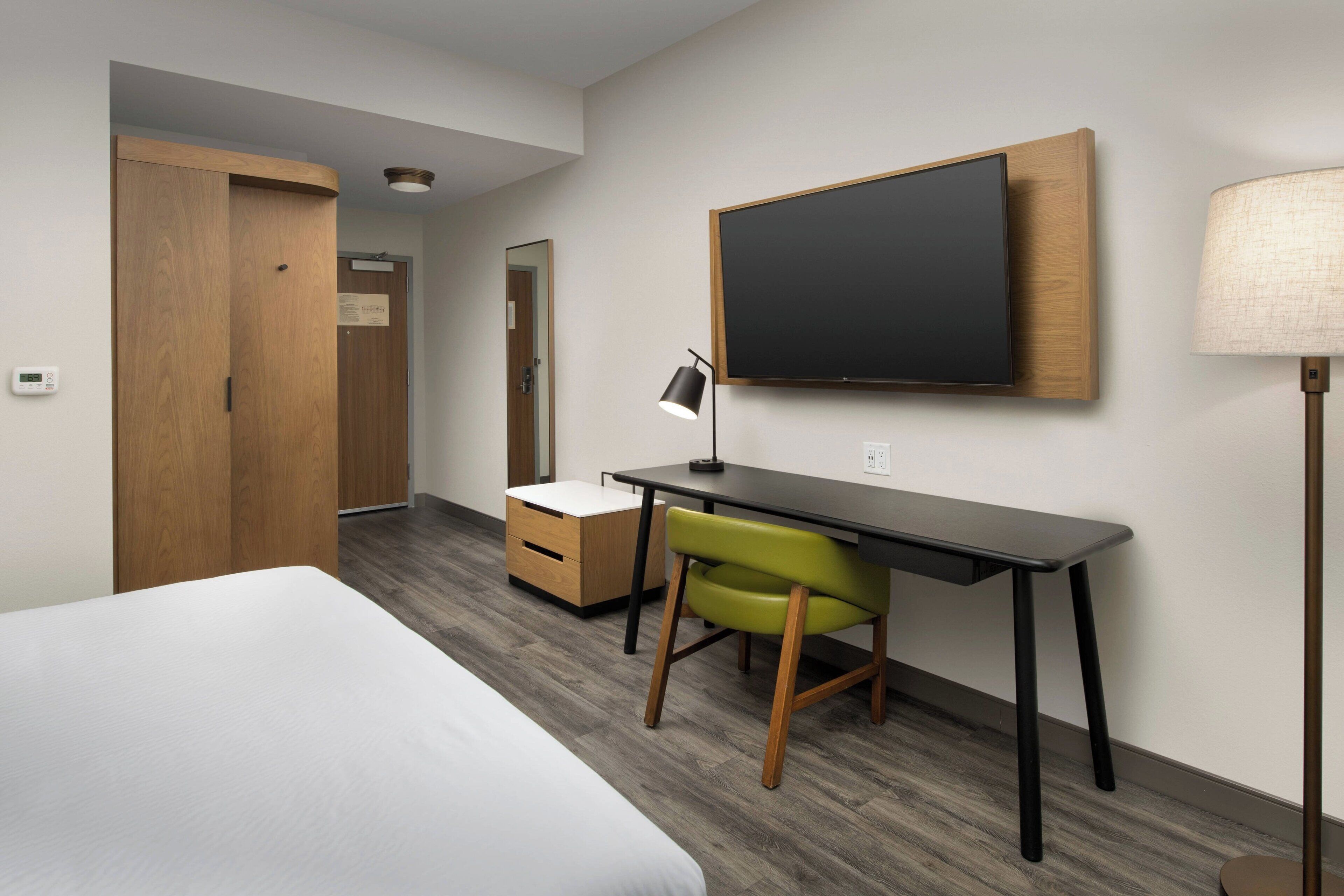 A hotel room with a bed, a desk with a chair, a flat-screen TV, a lamp, and a wardrobe.