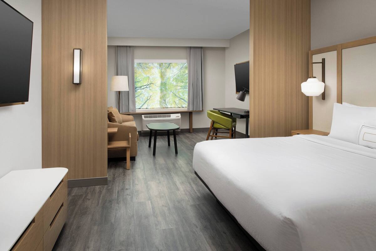 A hotel room with a large bed, a desk, a chair, a TV, and a window with a view of trees.