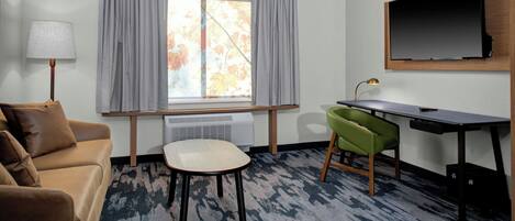 Une chambre d’hôtel avec un canapé, une petite table, un bureau, une chaise, une télévision et une fenêtre avec des rideaux.
