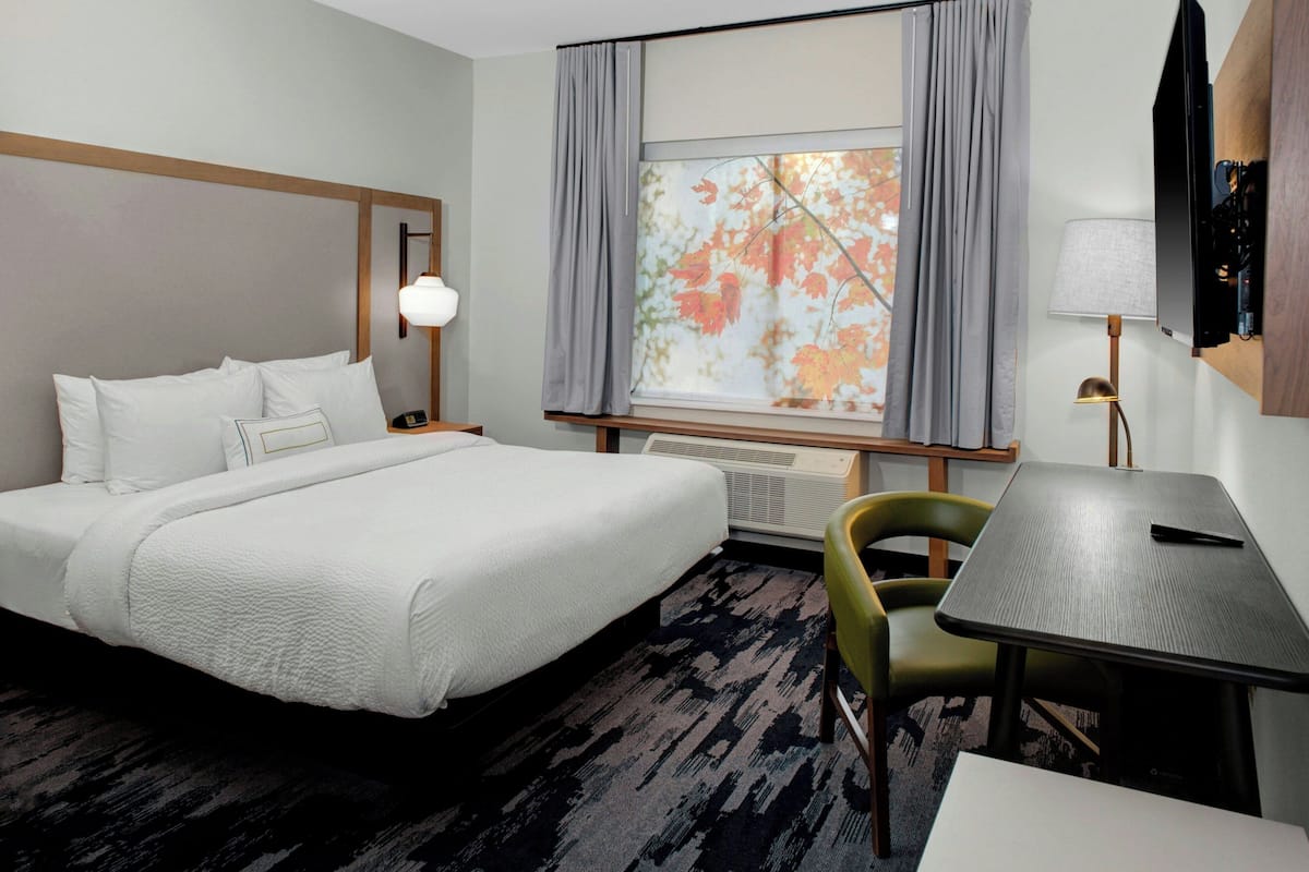 Een hotelkamer met een bed, bureau, stoel, raam met gordijnen, lamp en een flatscreen televisie aan de muur.