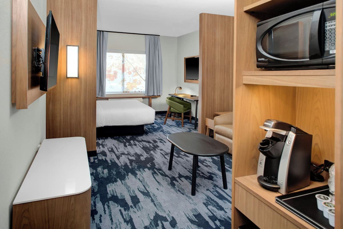 Een kleine hotelkamer met een bed, een bureau, een stoel, een koffiezetapparaat, een televisie en een raam met gordijnen.