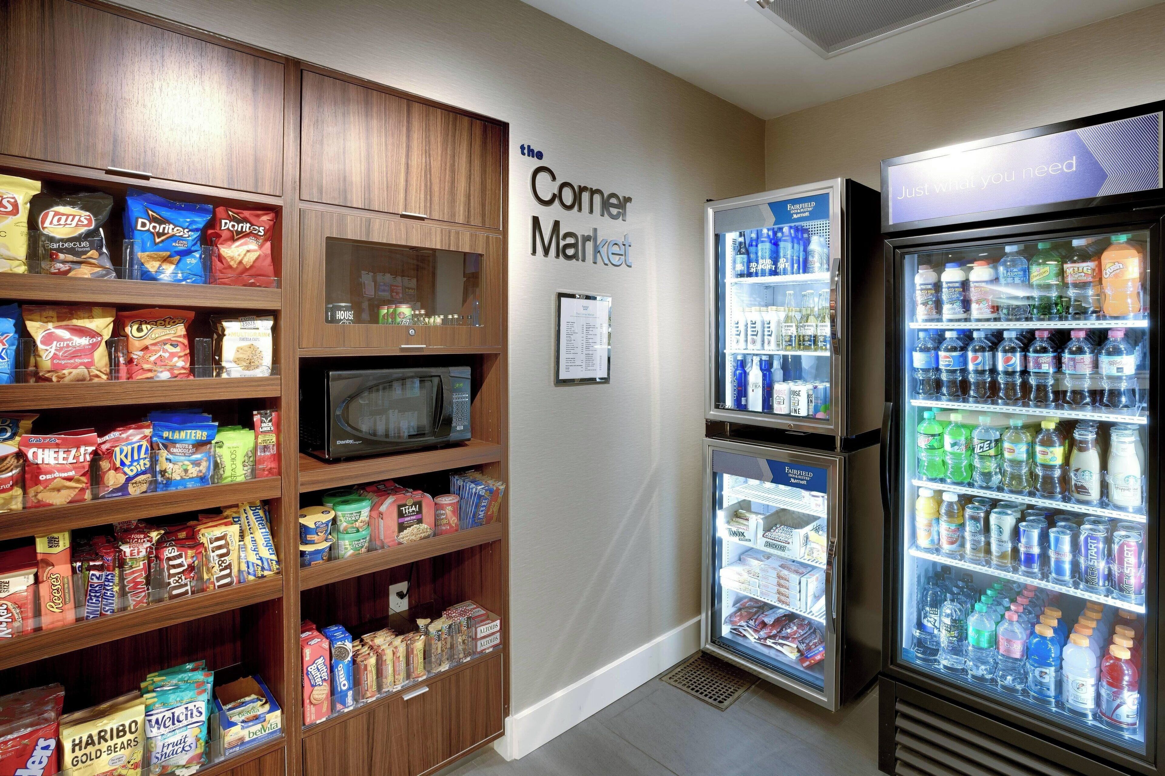 A corner market with shelves stocked with snacks, a microwave, and a refrigerator with beverages.