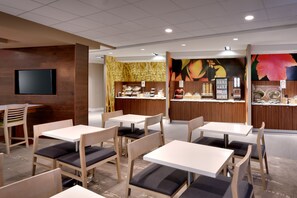 A modern restaurant interior with wooden tables and chairs, a flat-screen TV mounted on the wall, and a counter with a menu display.