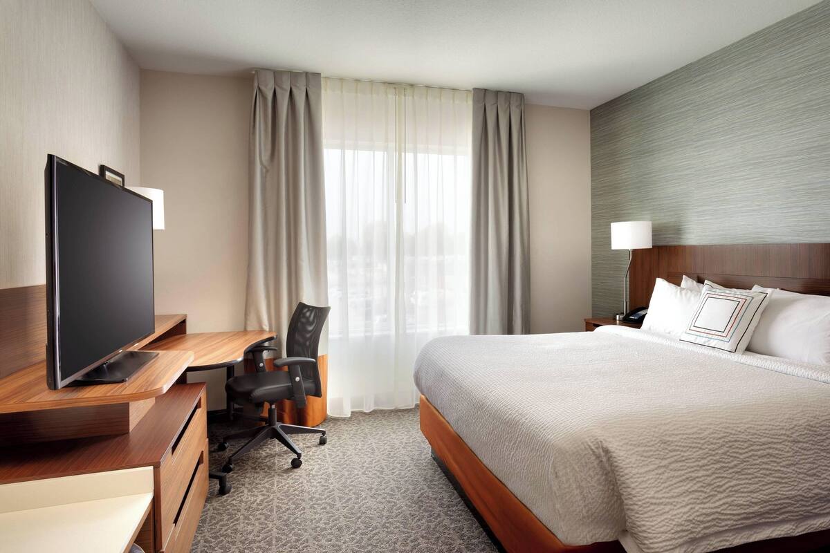 A hotel room with a bed, a desk with a TV, a chair, and a window with curtains.
