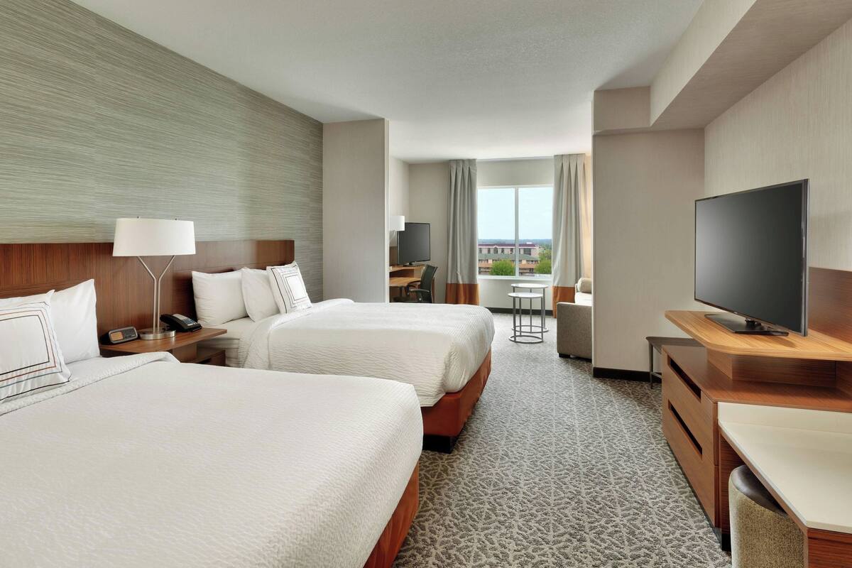 A hotel room with two beds, a desk, a TV, and a view of the city.