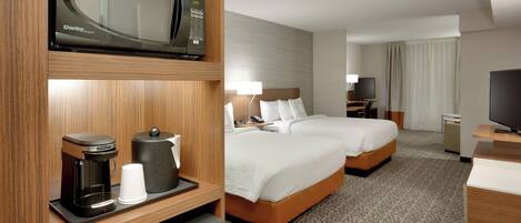 A hotel room with a microwave, coffee maker, and a TV on a wooden shelf. There are two beds with white linens and a desk with a lamp.
