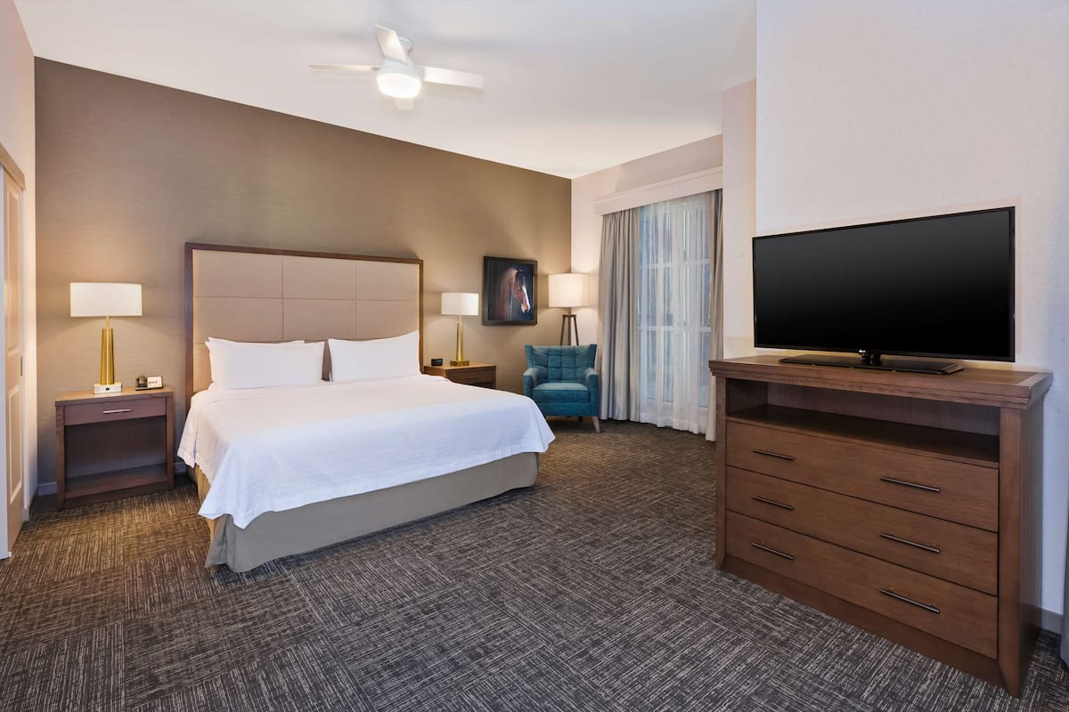 Quarto de hotel com uma cama grande, duas mesas de cabeceira com luminárias, uma TV de tela plana em um suporte de madeira, um ventilador de teto e uma janela com cortinas.