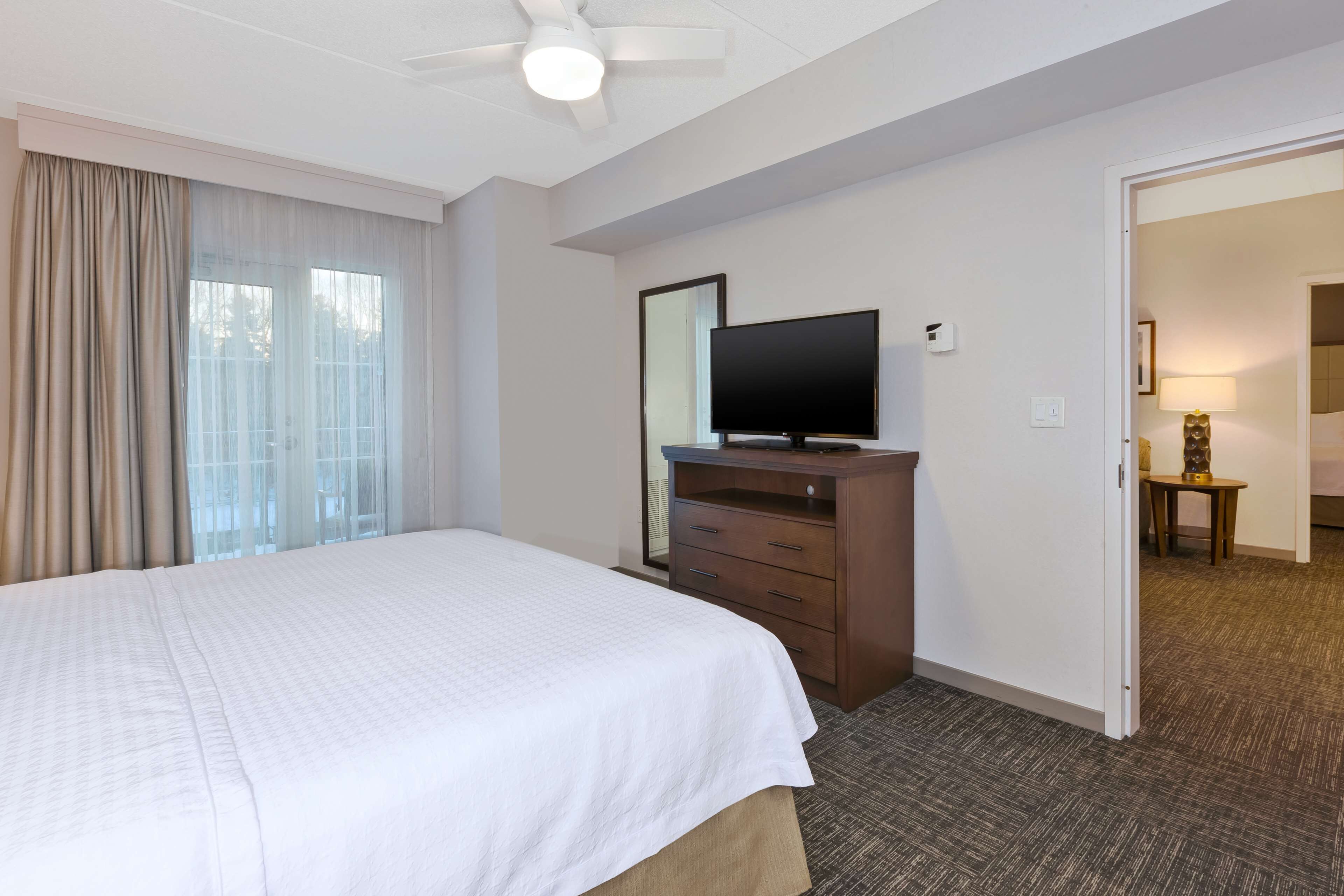 A hotel room with a large bed, a flat-screen TV on a wooden dresser, a ceiling fan, and a view of the outdoors through a window with curtains.