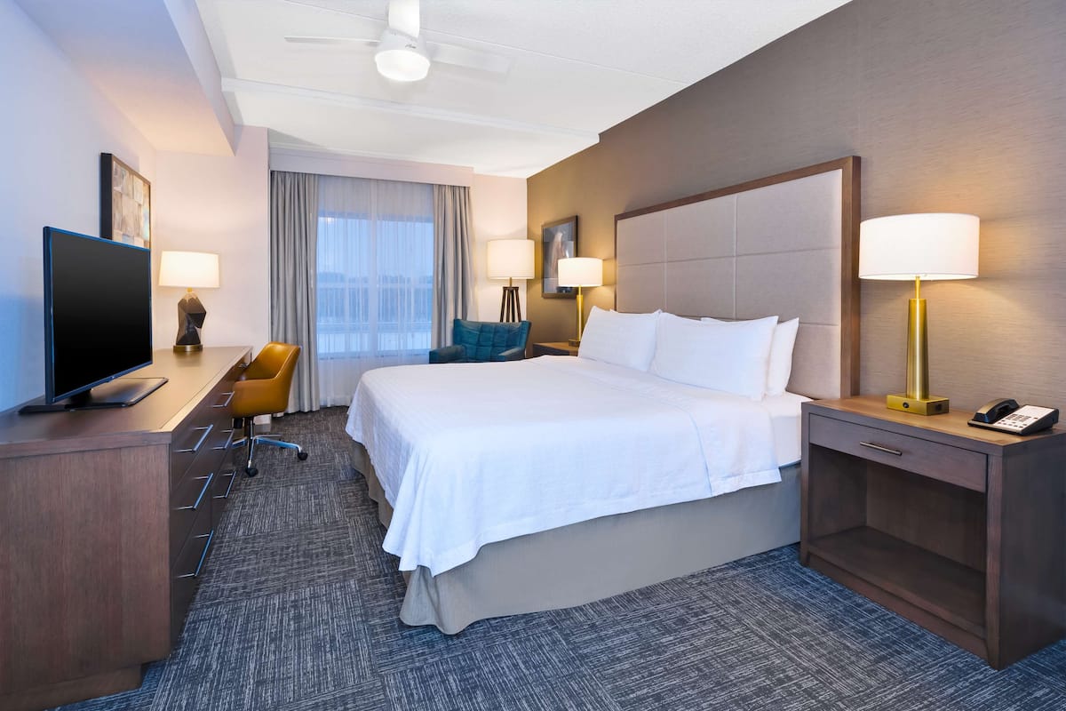 Quarto de hotel com uma cama grande, uma escrivaninha com televisão, duas luminárias, uma cadeira e uma janela com cortinas.