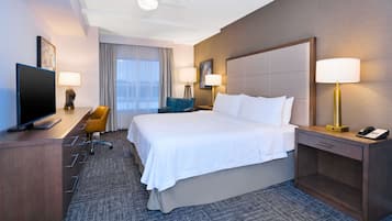 Habitación de hotel con una cama grande, un escritorio con televisor, dos lámparas, una silla y un ventanal con cortinas.