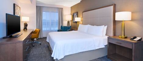 Habitación de hotel con una cama grande, un escritorio con televisor, dos lámparas, una silla y un ventanal con cortinas.