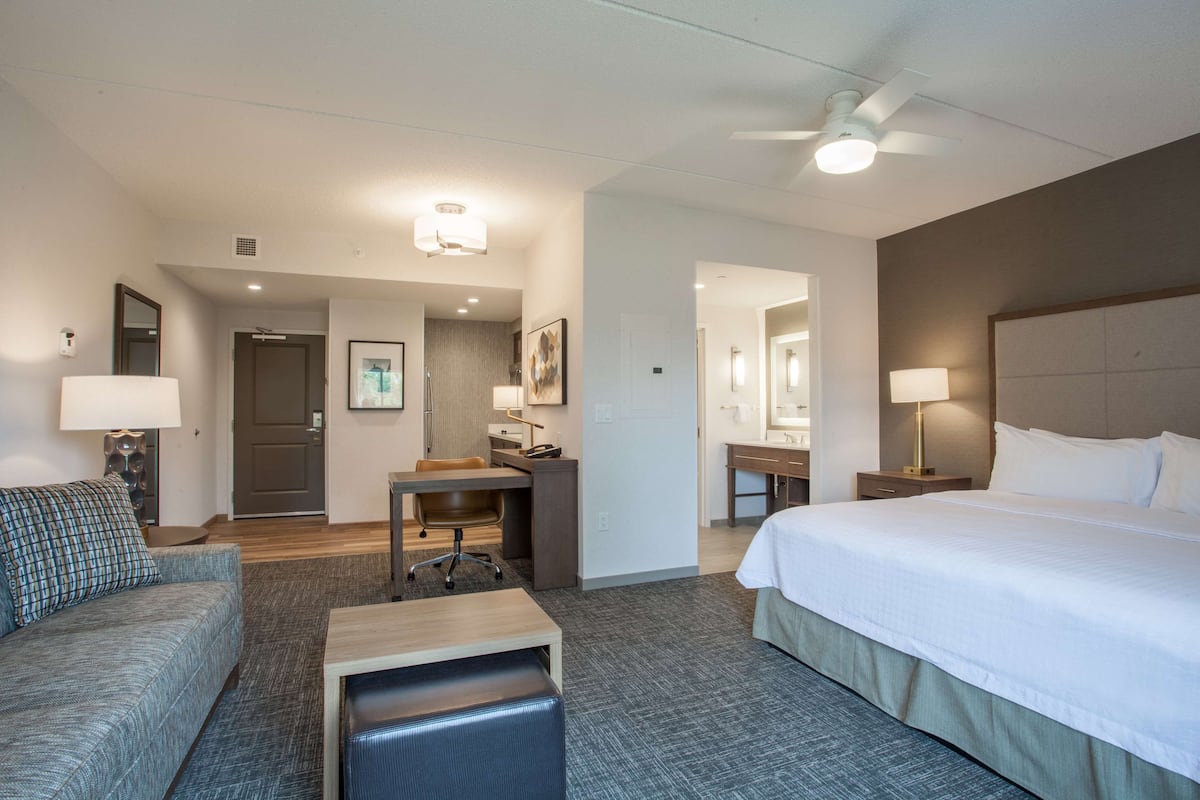 Quarto de hotel com cama, escrivaninha, poltrona, sofá, abajur e ventoinha de teto.
