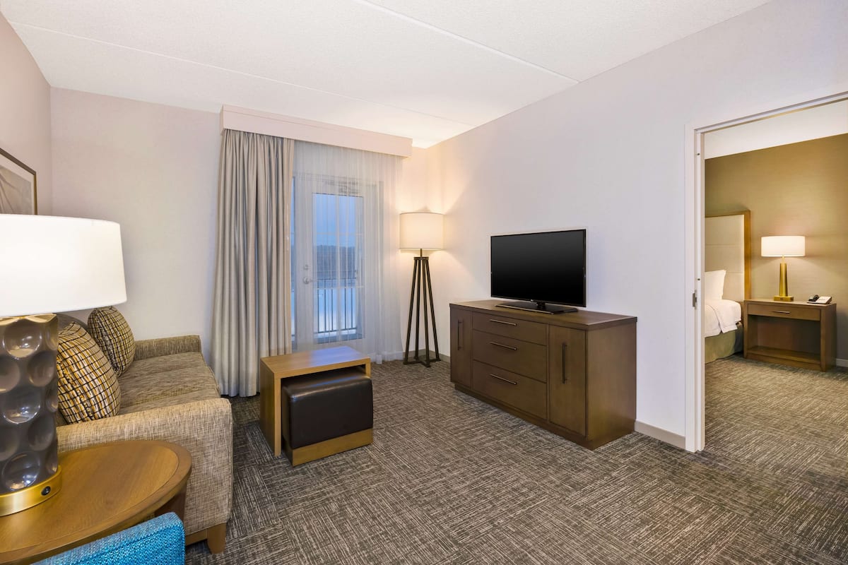 Quarto de hotel com sofá, televisão, secretária, candeeiro e vista para a cidade pela janela.