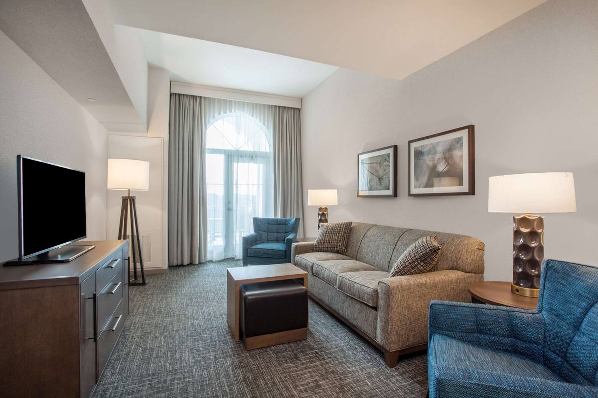 Quarto de hotel com televisão de tela plana, duas luminárias, um sofá, uma poltrona e um pequeno banquinho.