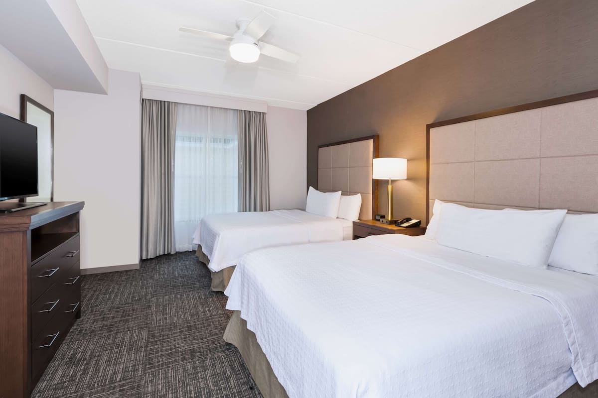 Quarto de hotel com duas camas, televisão, escrivaninha, abajur e ventoinha no teto.
