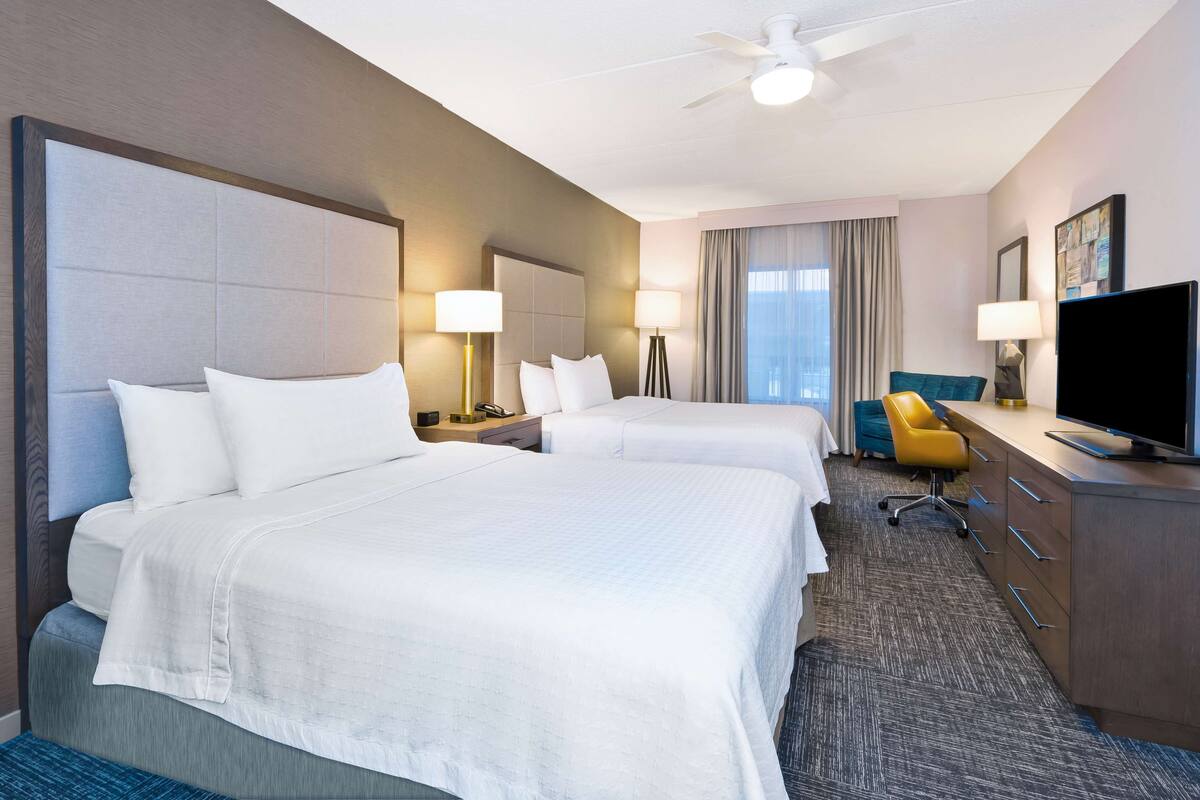 Quarto de hotel com duas camas, uma mesa de trabalho, televisão de tela plana, uma cadeira e um ventilador de teto.