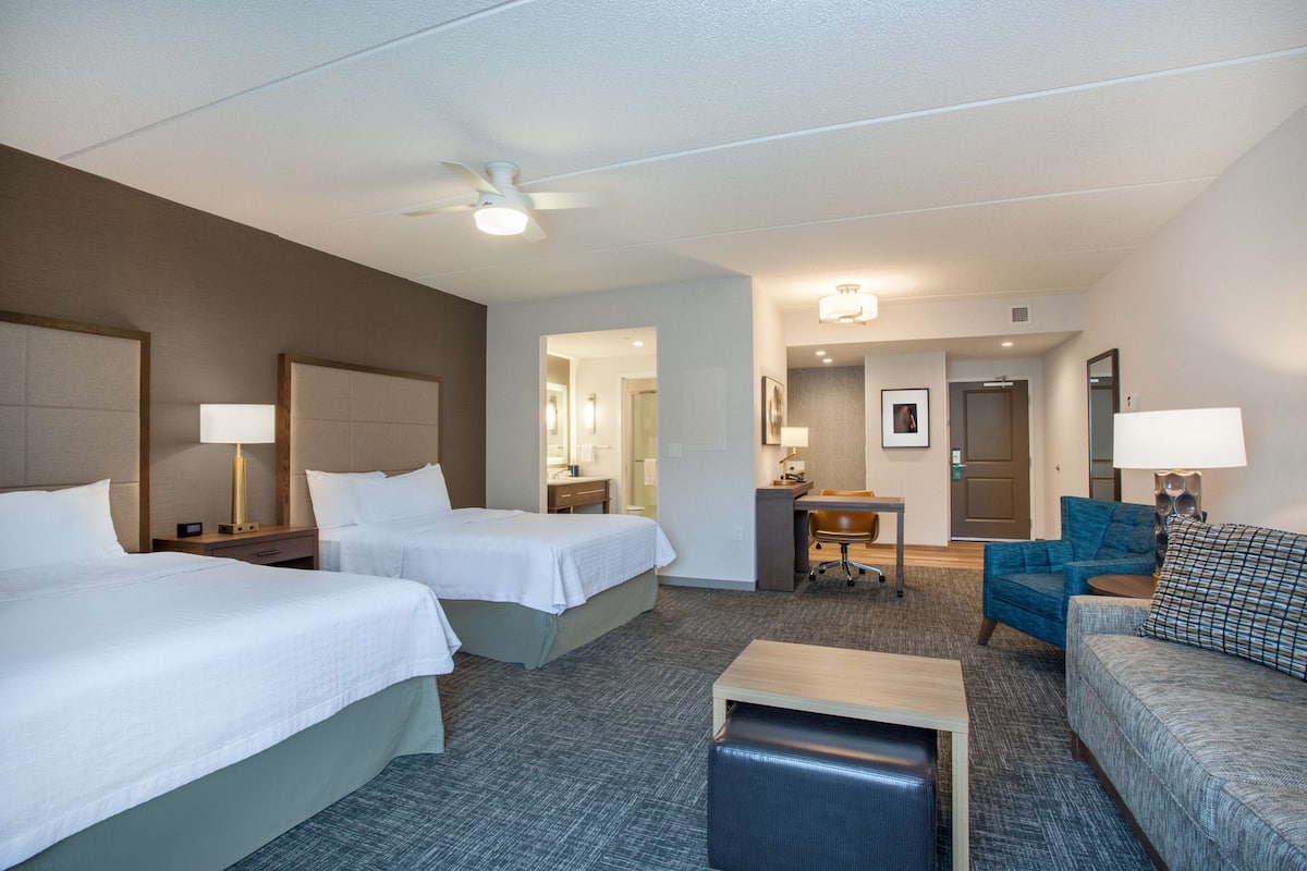 Quarto de hotel com duas camas, uma escrivaninha, uma cadeira, um sofá e um ventilador de teto.