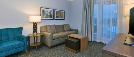 A hotel room with a blue sofa, a grey sofa, a small wooden table, a TV on a wooden stand, and two framed pictures on the wall.